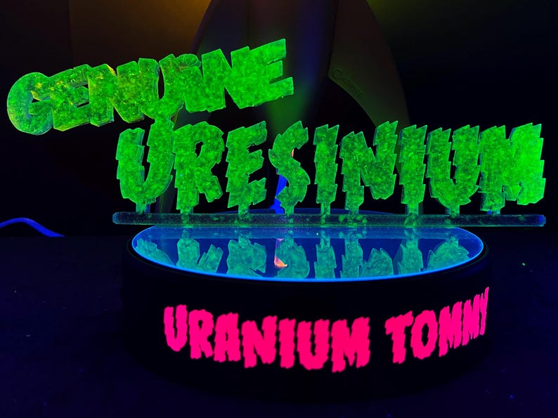 May include: A neon green sign that reads "GENUINE URESINIUM" above a black circular base with the words "URANIUM TOMMY" in pink. The sign and base are illuminated with a black light, creating a glowing effect.