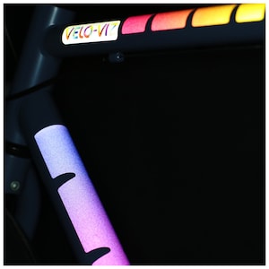 May include: A close-up of a bicycle frame with a reflective rainbow sticker that says "VELO-VIZ" on it.