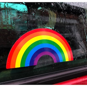 Könnte beinhalten: Ein Regenbogen-Aufkleber auf einem Autofenster mit Regentropfen. Der Aufkleber ist ein bunter Bogen mit roten, orangen, gelben, grünen, blauen, indigoblauen und violetten Streifen.