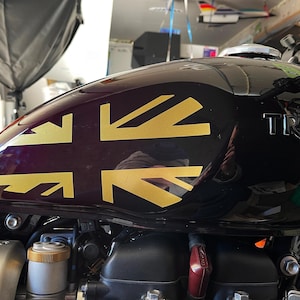 Puede incluir: Un tanque de gasolina de motocicleta negro con una calcoman&iacute;a Union Jack dorada y la palabra "TRIUMPH" en relieve en el lateral.