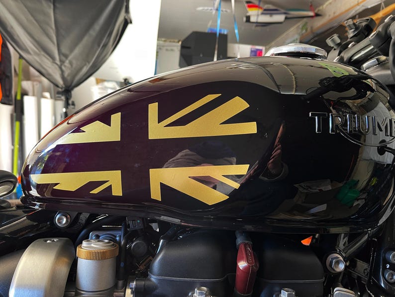 Puede incluir: Un tanque de gasolina de motocicleta negro con una calcoman&iacute;a Union Jack dorada y la palabra "TRIUMPH" en relieve en el lateral.