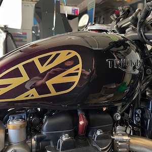 Puede incluir: Un tanque de gasolina de motocicleta negro con una calcoman&iacute;a Union Jack dorada y la palabra "TRIUMPH" en relieve en el lateral.