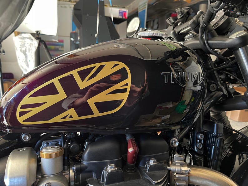 Puede incluir: Un tanque de gasolina de motocicleta negro con una calcoman&iacute;a Union Jack dorada y la palabra "TRIUMPH" en relieve en el lateral.