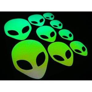 May include: A collection of glowing green alien head stickers on a black background. The stickers vary in size and have large, oval eyes. The color gradient shifts from a bright green to a lighter, almost white, hue at the bottom of each head.