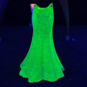 May include: A glowing green glass dress figurine. The dress has a fitted bodice and a flared skirt. The glass has a textured surface and is illuminated with a bright green light, creating a striking visual effect. The dress is a decorative item.