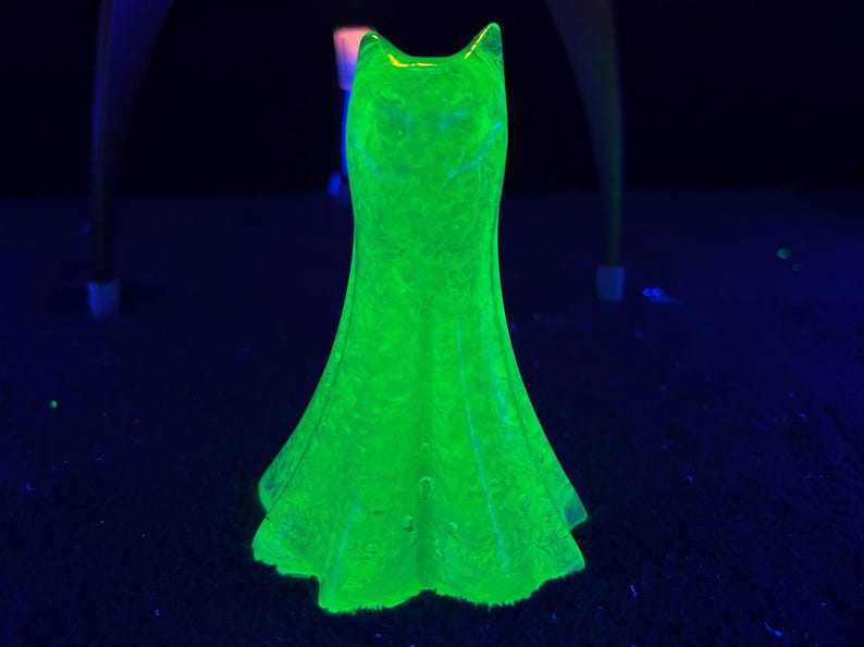 May include: A glowing green glass dress figurine. The dress has a fitted bodice and a flared skirt. The glass has a textured surface and is illuminated with a bright green light, creating a striking visual effect. The dress is a decorative item.