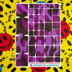 Puede incluir: Una hoja de pegatinas de camuflaje de color morado y negro para personalizar un teclado. Las pegatinas est&aacute;n dispuestas en una cuadr&iacute;cula y incluyen teclas de flecha, teclas de funci&oacute;n y otras teclas. El texto en la hoja dice "WE PRINT CUSTOM STICKERS! EMAIL US: HELLO@THEJOYBOX.CO.UK JOYBOXBUDE JOYBOXPRINTING".