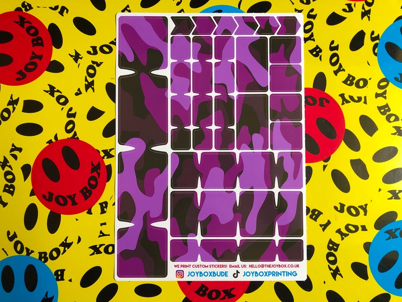 Puede incluir: Una hoja de pegatinas de camuflaje de color morado y negro para personalizar un teclado. Las pegatinas est&aacute;n dispuestas en una cuadr&iacute;cula y incluyen teclas de flecha, teclas de funci&oacute;n y otras teclas. El texto en la hoja dice "WE PRINT CUSTOM STICKERS! EMAIL US: HELLO@THEJOYBOX.CO.UK JOYBOXBUDE JOYBOXPRINTING".
