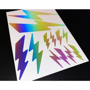 Könnte beinhalten: Bogen mit holografischen Aufklebern mit verschiedenen Blitzdesigns. Die Aufkleber wechseln die Farben von Lila zu Grün und Gold. Die Aufkleber befinden sich auf einem weißen Hintergrund.