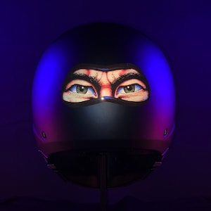 Könnte beinhalten: Ein schwarzer Motorradhelm mit einem aufgedruckten Bild von einem Paar Augen, die hinter einer schwarzen Maske hervorblicken.