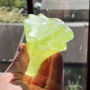 May include: A hand holding a translucent, lime green glass object. The object has a unique, organic shape with a flared top and a stem. The glass catches the light, creating a shimmering effect. The background shows a window and rooftops.