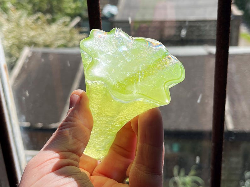 May include: A hand holding a translucent, lime green glass object. The object has a unique, organic shape with a flared top and a stem. The glass catches the light, creating a shimmering effect. The background shows a window and rooftops.
