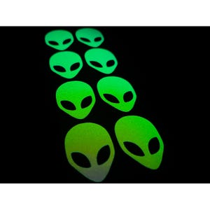 Puede incluir: Pegatinas de cabeza de alienígena que brillan en la oscuridad en varios tamaños. Las pegatinas son de color verde neón brillante con ojos ovalados negros y una superficie texturizada. Las pegatinas están dispuestas en un patrón diagonal sobre un fondo negro.
