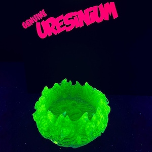 May include: A small, circular, translucent green bowl with a jagged, flame-like rim. The object glows intensely under ultraviolet light. Above the bowl, the text "GENUINE URESINIUM" is printed in bright pink, bold, sans-serif capital letters.