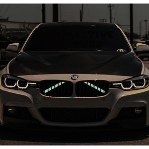 Op de afbeelding: Een witte BMW sedan met een zwarte motorkap en een zwarte grille. De auto heeft zwarte koplampen en een zwarte bumper. De auto staat 's nachts op straat geparkeerd.