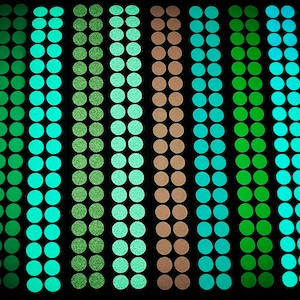 May include: A close-up of seven rows of glow-in-the-dark round stickers in various shades of green and blue. The stickers are arranged in vertical columns.