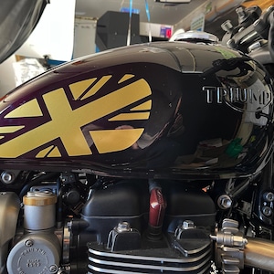 Puede incluir: Un tanque de gasolina de motocicleta negro con una calcoman&iacute;a Union Jack dorada y la palabra "TRIUMPH" en relieve en el lateral.