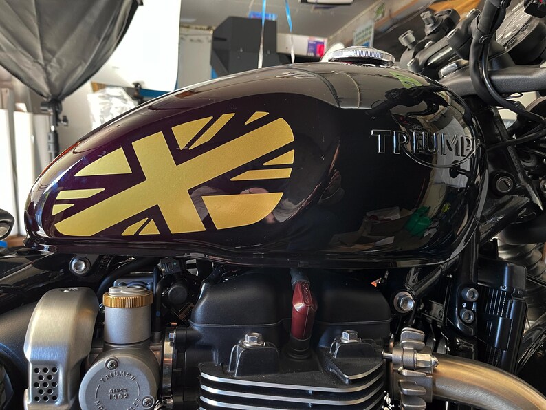 Puede incluir: Un tanque de gasolina de motocicleta negro con una calcoman&iacute;a Union Jack dorada y la palabra "TRIUMPH" en relieve en el lateral.