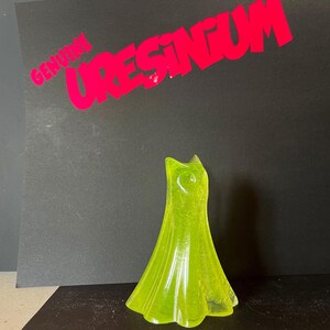 May include: A translucent, lime green dress-shaped figurine. The figurine is standing upright against a black background. The text "GENUINE URESINGUM" is printed in red above the figurine.