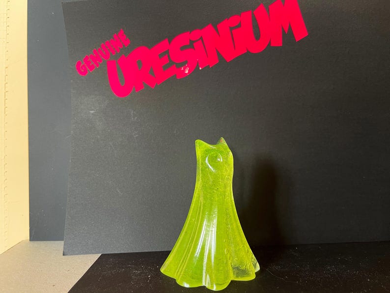 May include: A translucent, lime green dress-shaped figurine. The figurine is standing upright against a black background. The text "GENUINE URESINGUM" is printed in red above the figurine.