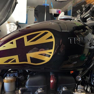 Puede incluir: Un tanque de gasolina de motocicleta negro con una calcoman&iacute;a Union Jack dorada y la palabra "TRIUMPH" en relieve en el lateral.
