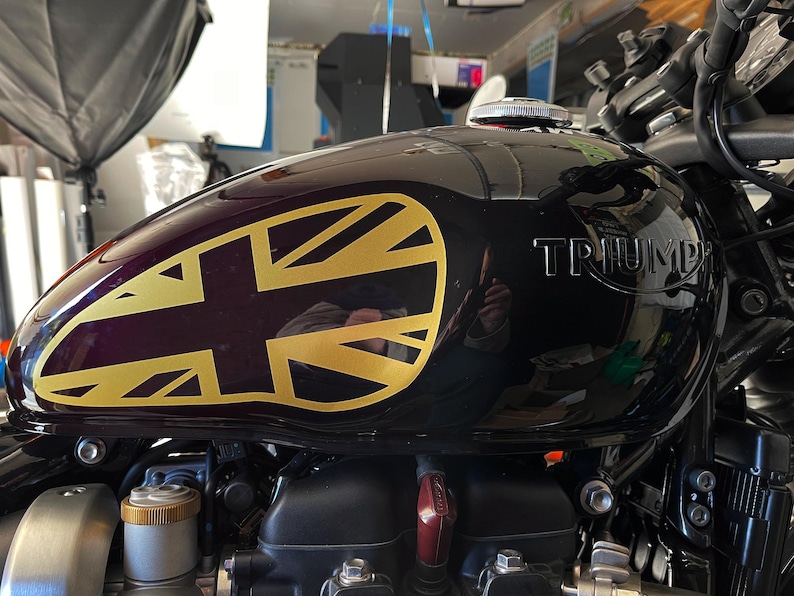 Puede incluir: Un tanque de gasolina de motocicleta negro con una calcoman&iacute;a Union Jack dorada y la palabra "TRIUMPH" en relieve en el lateral.