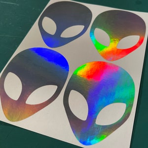 Peut inclure: Quatre autocollants tête d'extraterrestre holographiques irisés sur une feuille de papier blanche. Les autocollants sont un arc-en-ciel de couleurs, y compris le bleu, le vert, le rouge et l'orange.