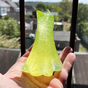 May include: A translucent, lime green glass dress figurine. The dress has a textured surface and a flared skirt. The figurine is held in front of a window with a blurred background of houses and trees.