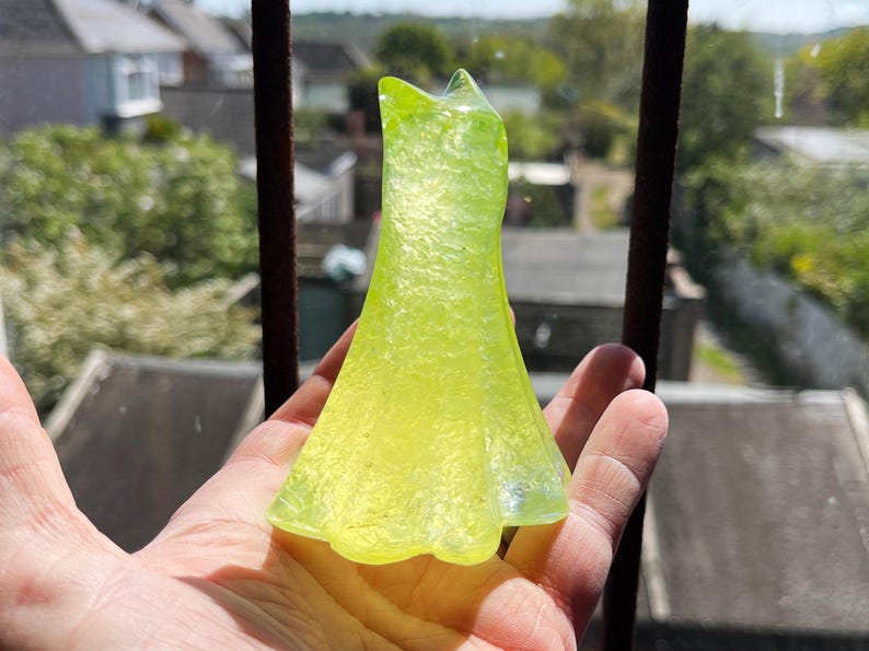 May include: A translucent, lime green glass dress figurine. The dress has a textured surface and a flared skirt. The figurine is held in front of a window with a blurred background of houses and trees.