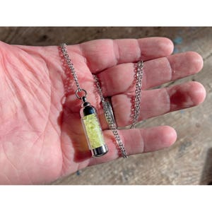 May include: A silver necklace with a pendant. The pendant is a small, clear vial filled with light green crystals. The vial has silver caps and hangs from a delicate chain. The necklace is displayed against a neutral background.