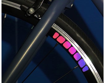May include: A close-up of a bicycle wheel with a pink and purple reflective stripe. The stripe is made up of individual squares that fade from pink to purple.