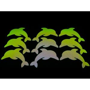 以下が含まれることがあります： 黒い背景に、緑と銀色の9つのイルカ型ステッカー。ステッカーはキラキラと反射する仕上がりで、きらめく効果を生み出しています。イルカはパターン状に配置されています。