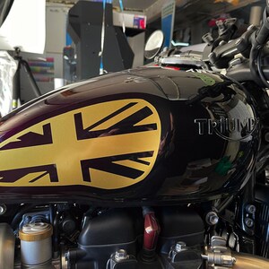 Puede incluir: Un tanque de gasolina de motocicleta negro con una calcoman&iacute;a de la Union Jack dorada. El tanque tiene la palabra "TRIUMPH" impresa en &eacute;l.