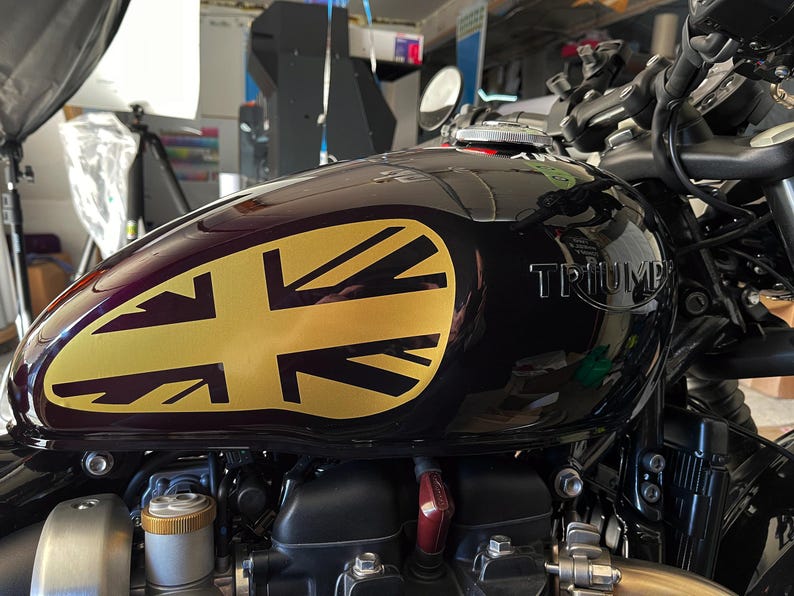 Puede incluir: Un tanque de gasolina de motocicleta negro con una calcoman&iacute;a de la Union Jack dorada. El tanque tiene la palabra "TRIUMPH" impresa en &eacute;l.