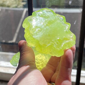 May include: A translucent, lime green, abstract glass sculpture with a textured surface. The art piece is held up in front of a window, catching the sunlight. The sculpture has a unique, organic shape.