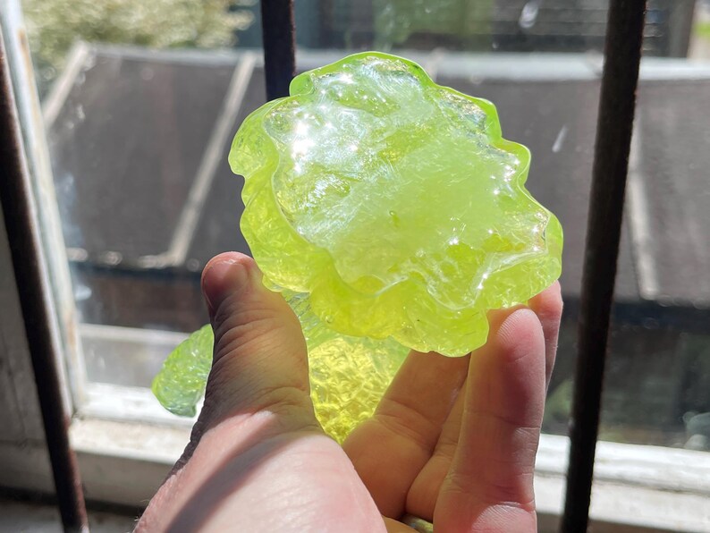 May include: A translucent, lime green, abstract glass sculpture with a textured surface. The art piece is held up in front of a window, catching the sunlight. The sculpture has a unique, organic shape.