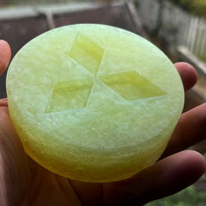 Può includere: Un oggetto circolare, traslucido e giallo pallido con una superficie ruvida e leggermente bollicinosa. Al centro della faccia superiore è impresso un logo a tre diamanti, caratterizzato da tre rombi identici disposti a triangolo.