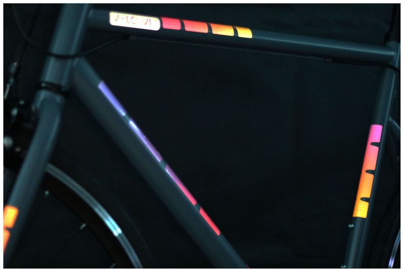 May include: A black bicycle frame with colorful reflective tape accents. The tape is in a rainbow pattern with red, orange, yellow, green, blue, indigo, and violet colors. The tape is on the top tube and down tube of the frame.