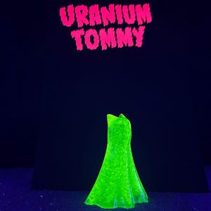 May include: A neon green dress is displayed against a dark blue background. The dress is illuminated, showcasing its shape and texture. Above the dress, the words "URANIUM TOMMY" are written in bright pink, adding a bold contrast.