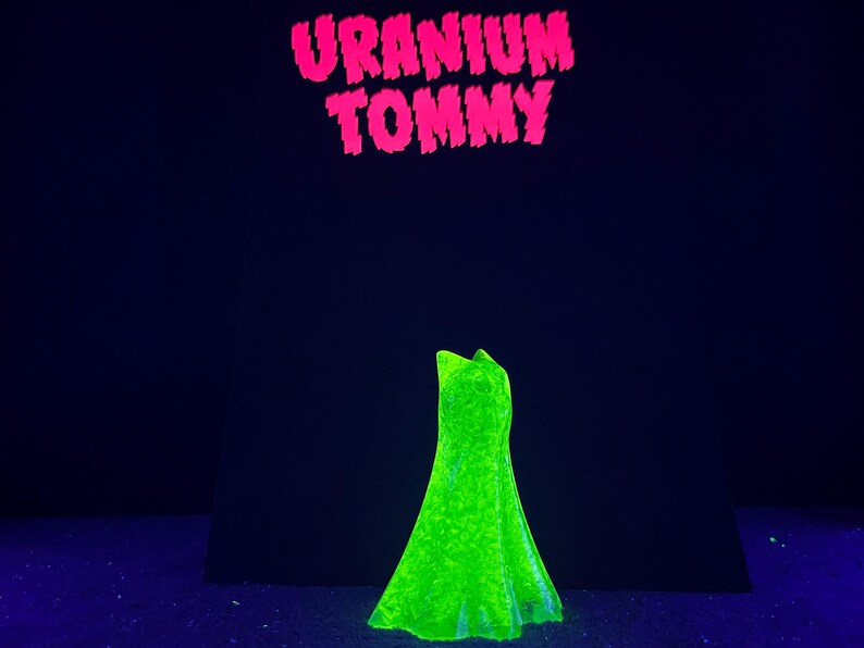 May include: A neon green dress is displayed against a dark blue background. The dress is illuminated, showcasing its shape and texture. Above the dress, the words "URANIUM TOMMY" are written in bright pink, adding a bold contrast.