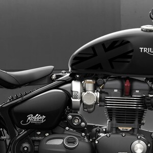 Puede incluir: Una motocicleta Triumph Bonneville Bobber negra con un dise&ntilde;o de Union Jack en el tanque de gasolina. La motocicleta tiene un asiento de cuero negro y un motor cromado.