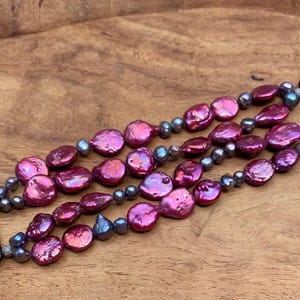 May include: A three-strand watch band featuring large, iridescent, pinkish-red freshwater pearls and smaller, gray-blue beads. The band has black connectors for the watch and rests on a wooden surface.