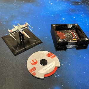 May include: A black plastic storage box with a clear plastic insert holds a stack of cards with the text "Rebel Alliance" and "T-65 X-wing". A small, white and red painted model of a T-65 X-wing spaceship sits on a black base. A round, gray and red token with the text "T-65 X-wing" is on a blue surface.