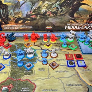 以下が含まれることがあります： 「Epic Battles in the Lands of Middle-Earth」というボードゲーム。詳細な地図上に、さまざまなミニチュアフィギュアとゲームの駒が配置されています。ゲームの駒は赤、青、白です。ゲームの箱が背景に見えます。