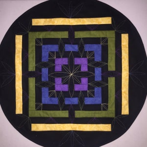 May include: A round black quilted table runner with a geometric design in yellow, green, blue, and purple. The design features a central square with a flower-like pattern surrounded by concentric squares.