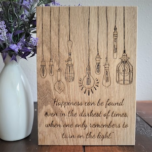 May include: A wooden sign with a quote about finding happiness in the darkest of times. The quote is "Happiness can be found even in the darkest of times, when one only remembers to turn on the light." The sign is decorated with a drawing of light bulbs hanging from the ceiling.