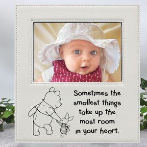 Könnte beinhalten: Weißer Fotorahmen mit einem weißen Passepartout und einem Bild eines Babys, das einen weißen Sonnenhut trägt. Der Rahmen hat ein Winnie Puuh-Zitat, das lautet: "Manchmal nehmen die kleinsten Dinge den meisten Platz in deinem Herzen ein."