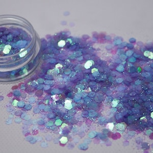 May include: A small jar of iridescent blue and purple glitter. The glitter is scattered around the jar on a white surface.