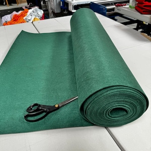 Puede incluir: Un rollo de tela de fieltro verde oscuro se desenrolla sobre una superficie blanca. Un par de tijeras negras est&aacute; sobre la tela.
