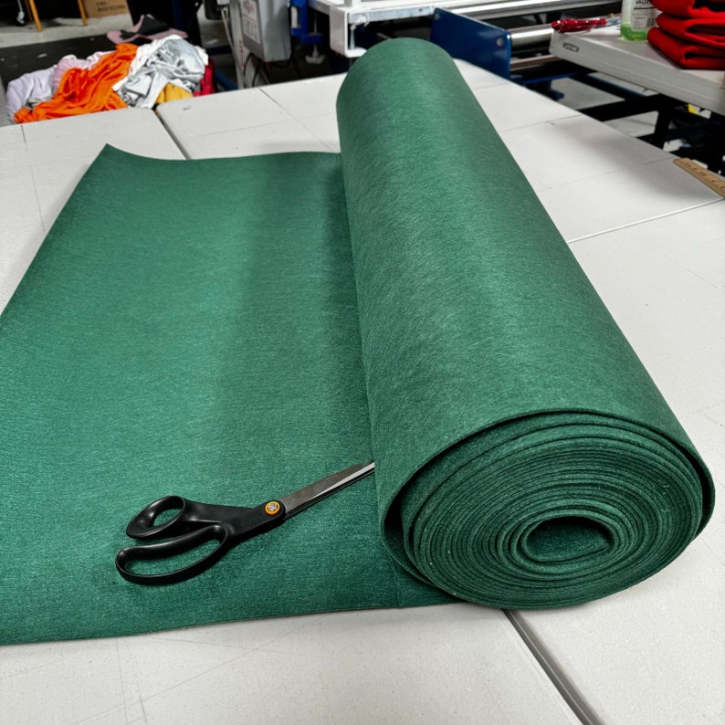 Puede incluir: Un rollo de tela de fieltro verde oscuro se desenrolla sobre una superficie blanca. Un par de tijeras negras est&aacute; sobre la tela.